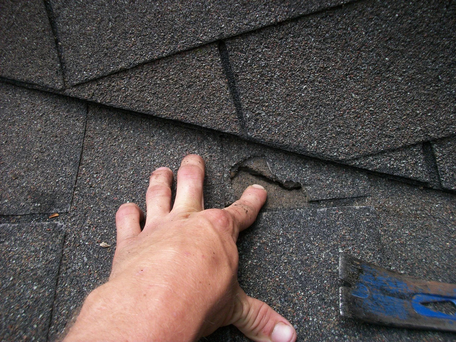 A hand lifts a damaged asphalt shingle beside a blue pry bar during a localized roof repair
